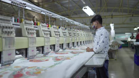 Joven-Indio-Con-Mascarilla-Trabajando-En-Una-Fábrica-Textil-Revisando-El-Trabajo-De-Una-Máquina-De-Coser-Moderna---Bangalore,-India