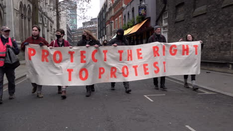 Menschen-Marschieren-Entlang-Einer-Straße-Mit-Einem-Weißen-Banner-Mit-Orangefarbener-Aufschrift,-Auf-Dem-Während-Eines-Kill-the-Bill-Protestes-„Schutz-Des-Rechts-Auf-Protest“-Steht