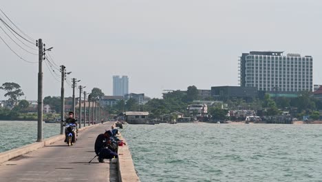 Un-Hombre-Preparando-Su-Caña-De-Pescar-Mientras-Pasa-Una-Motocicleta-Y-La-Ciudad-De-Pattaya-En-El-Fondo-En-El-Muelle-De-Pesca-De-Pattaya,-Chonburi,-Tailandia