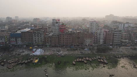 Un-Dron-Retrocede-Sobre-El-Río-En-El-Viejo-Dhaka-Mostrando-Los-Edificios-Altos-Y-Los-Barcos-Que-Bajan-Por-El-Río
