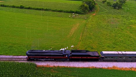 Una-Vista-Aérea-Paralela-De-Un-Antiguo-Tren-De-Pasajeros-De-Vapor-Que-Sopla-Humo-Negro-Bajo-La-Lluvia-A-Través-De-Las-Tierras-Agrícolas-De-Pensilvania