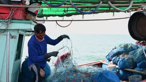 Reparando-La-Red-Mientras-Fuma-Y-Luego-Sonríe-A-La-Cámara-Mientras-El-Barco-Se-Mueve-Con-Las-Olas,-Muelle-De-Pesca-De-Pattaya,-Chonburi,-Tailandia