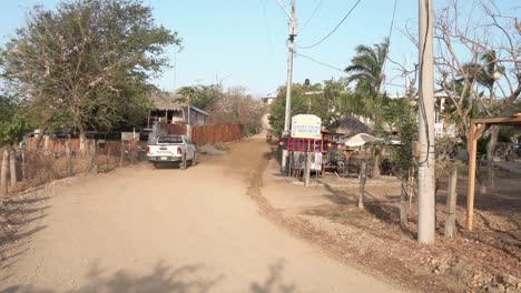 Man-and-woman-riding-a-motorcycle-pass-a-low-cost-hotel-on-a-dirt-road,-Handheld-stable-shot