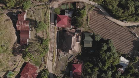 grass-roof-house-hut-in-a-mountain-village-red-tops-of-homes-farms-in-backyard-with-trails-weaving-through-trees-connecting-community-aerial-rotating-ascending-wide-angle-birds-eye-view-top-down