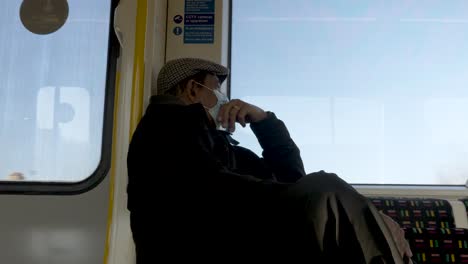 A-close-up-shot-of-a-passenger-wearing-a-face-mask-gazing-out-of-the-window-of-a-train,-the-man-on-his-daily-commute-along-the-Metropolitan-line-from-Amersham-to-Aldgate,-London,-England