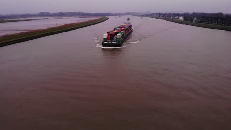 Antena-Sobre-El-Río-Noord-Con-Vista-Del-Barco-Fps-Rijn-Que-Transporta-Contenedores-Acercándose-En-Un-Día-Nublado