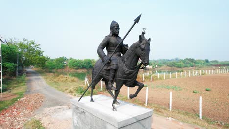 Black-statue-of-a-warrior-with-spear,-helmet-and-horse-going-forward-located-near-a-green-field-and-yellow-step-in-summer-of-Vadodara,-India