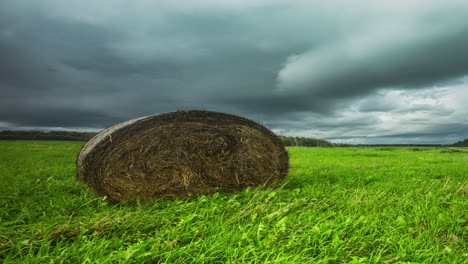 Low-angle-shot-of-dark-rain-clouds-passing-by-over-green-grass-field-with-a-hay-bale-in-timelapse-in-the-evening-sky