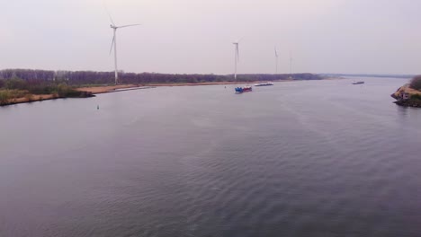 Vista-Aérea-Sobre-Oude-Maas-En-Un-Día-Nublado-Con-Turbinas-Eólicas-Inmóviles-En-El-Borde-De-Los-Ríos-Y-Buques-De-Carga-Acercándose-En-La-Distancia