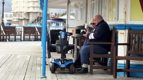 Pareja-De-Ancianos-Jubilados-Con-Scooter-De-Movilidad-Relajándose-En-El-Paseo-Marítimo-Del-Muelle-Galés-De-Estancia