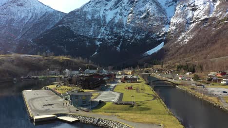 Hermosa-Flam-Noruega---Antena-En-Movimiento-Hacia-Atrás-Sobre-Un-Fiordo-Claro-Con-Reflejos-De-Sol-Y-Montaña---Desde-El-Cierre-De-La-Ciudad-Y-El-Muelle-De-Cruceros-Hasta-Una-Vista-Panorámica-Con-Fondo-De-Montaña