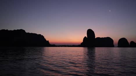 Boot,-Das-Auf-Dem-Glasigen-Wasser-Während-Eines-Orangefarbenen-Sonnenaufgangs-Am-Morgen-In-Krabi,-Thailand,-Fährt,-Mit-Silhouetten-Von-Kalksteinbergen-In-Der-Ferne