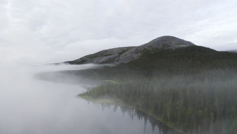 Aerial-view-of-flying-up-and-above-mist-over-a-lake-revealing-a-forest-and-mountain-landscape