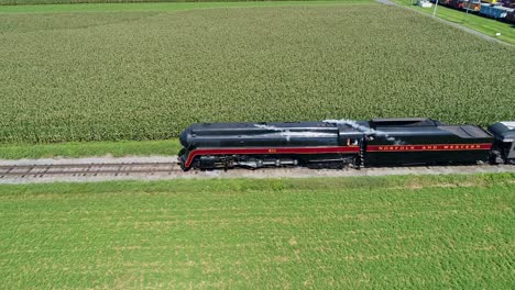 Una-Vista-Aérea-Que-Viaja-Al-Costado-De-Un-Antiguo-Tren-De-Pasajeros-De-Vapor,-Que-Sopla-Humo-Que-Viaja-A-Través-De-Campos-De-Maíz-Y-Ricas-Tierras-De-Cultivo-En-Un-Día-De-Verano-Sin-Nubes