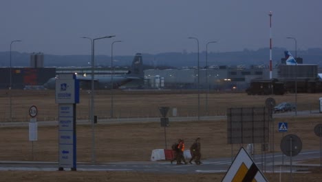 Un-Avión-De-Carga-Militar-Espera-El-Despegue-En-La-Distancia,-Cuatro-Miembros-Del-Personal-De-Seguridad-Del-Aeropuerto-Cruzan-Una-Carretera