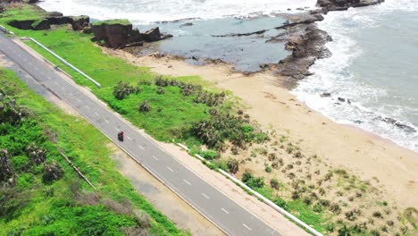 Luftdrohnenansicht-Von-Zwei-Personen-Auf-Einem-Zweirad,-Die-Auf-Einer-Gut-Asphaltierten,-Leeren-Straße-Mit-Einem-Sandstrand-Auf-Der-Rechten-Seite-Fahren,-Weiße-Wellen,-Die-Sanft-Mit-Felsformationen-Und-Grün-Am-Ufer-Krachen