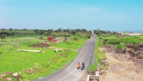 aerial-view-of-man-and-woman-on-two-wheelers-approaching-camera-from-the-other-direction,-bright-blue-clear-sky-in-background,-grass-on-sides-of-road,-carefree-couple,-focus-on-background,-scenic-view