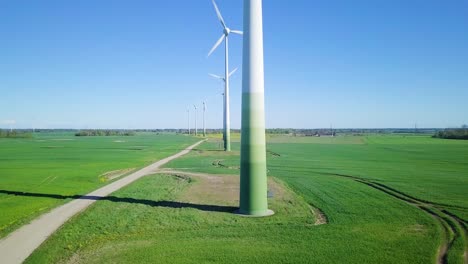 Aerial-view-of-wind-turbines-generating-renewable-energy-in-the-wind-farm,-sunny-spring-day,-low-flyover-over-green-agricultural-cereal-fields,-countryside-roads,-wide-angle-ascending-drone-shot