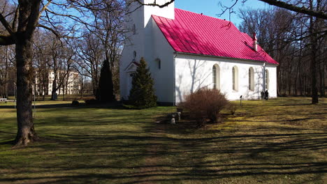 Aerial-view-of-Lielvarde-Lutheran-church-at-the-bank-of-Daugava-river,-white-church-with-red-roof,-leafless-trees,-sunny-spring-day,-low-angle-wide-shot-moving-forward