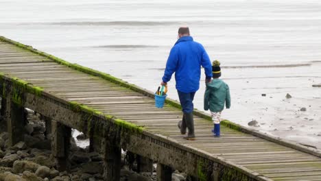 Padre-Sosteniendo-La-Cuchara-Junto-Al-Mar-Explorando-La-Marea-Baja-Embarcadero-Del-Paseo-Marítimo-De-La-Playa-Del-Reino-Unido-Con-Su-Hijo