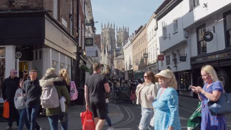 A-busy-street-scene-with-shoppers-and-tourists-in-the-ancient-medieval-city-of-York-on-a-bright-sunny-day-with-York-Minster-in-the-background-Yorkshire-England