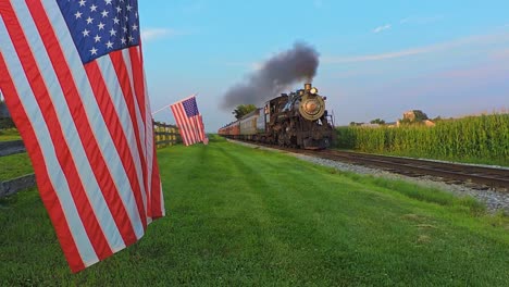 Una-Vista-De-Una-Línea-De-Banderas-Americanas-Que-Ondean-Suavemente-En-Una-Valla-Junto-A-Tierras-De-Cultivo-Mientras-Un-Tren-De-Pasajeros-De-Vapor-Que-Sopla-Humo-Se-Acerca-Durante-La-Hora-Dorada