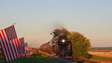 Una-Vista-De-Una-Línea-De-Banderas-Americanas-Que-Ondean-Suavemente-En-Una-Valla-Junto-A-Tierras-De-Cultivo-Mientras-Un-Tren-De-Pasajeros-De-Vapor-Que-Sopla-Humo-Se-Acerca-Durante-La-Hora-Dorada