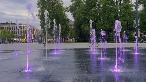 Colorful-ground-level-fountains-gushing-water-at-Lukiskes-square-at-sunset-in-Vilnius-and-children-having-fun-on-bicycles,-Lithuania