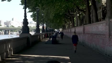 Gente-En-Público-Pasando-Su-Mañana-Caminando-Tranquilamente-Por-El-Sendero-Con-El-Río-Támesis-A-La-Izquierda-Y-El-Muro-Conmemorativo-Nacional-Covid-A-La-Derecha,-Londres,-Inglaterra