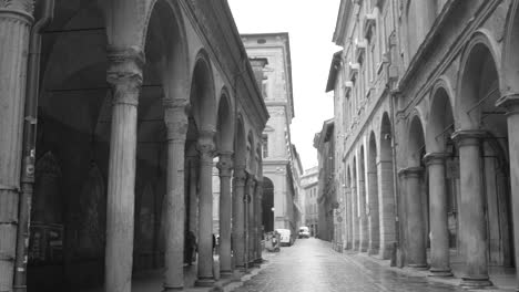 Fotografía-En-Blanco-Y-Negro-De-Gente-Caminando-Por-La-Acera-Bajo-Pórticos-En-Bolonia,-En-El-Norte-De-Italia,-Durante-El-Día