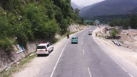 Aerial-drone-shot-over-cars-passing-by-along-the-winding-road-leading-upto-Manali,-Himachal-Pradesh,-India-along-the-mountain-slope-on-a-cloudy-day