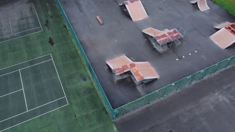 Aerial-Birdseye-view-flying-above-fenced-skate-park-ramp-in-empty-closed-playground-descending-tilt-up