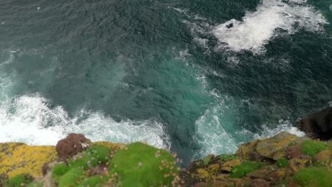 Waves-gently-crash-over-rocks-and-against-the-base-of-a-sea-cliff-in-the-deep-teal-coloured-ocean-while-seabirds-fly-around-the-cliffs-of-a-seabird-colony-of-guillemots-on-Handa-Island,-Scotland