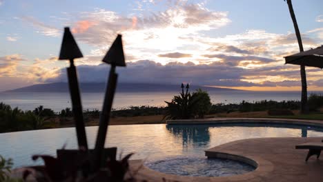 static-shot-of-pool-deck-view-of-sunset-behind-lanai-with-hot-tub-and-torches-on-in-maui-hawaii