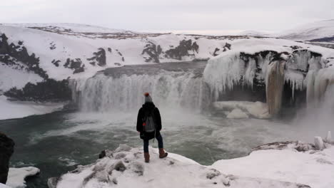 Aerial-Orbit-View-of-Tourist-Standing-at-the-Edge-of-Snowy-Cliff-at-Goðafoss-Waterfall-Iceland,-Revealing-Spectacular-Winter-Landscape-Around