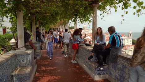 Turistas-Sentados-En-El-Borde-Del-Miradouro-De-Santa-Luiza-Acompañados-Por-Artistas-Que-Muestran-Sus-Ilustraciones-De-Lisboa-Y-Por-Músicos-Que-Brindan-Una-Banda-Sonora