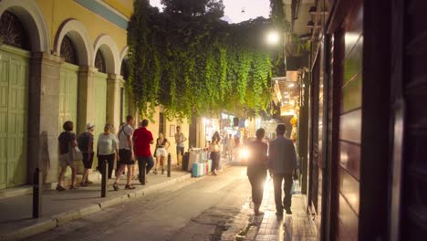 Leute,-Die-Eine-Schmale-Straße-In-Athen-Hinuntergehen,-Die-Von-Den-Scheinwerfern-Eines-Autos-Hinterleuchtet-Wird