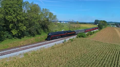 Una-Vista-Aérea-De-Un-Antiguo-Tren-De-Pasajeros-De-Vapor-Que-Se-Acerca-Soplando-Humo-Y-Vapor-Viajando-A-Través-De-Fértiles-Campos-De-Maíz-En-Un-Soleado-Día-De-Otoño