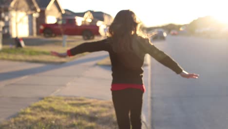 Little-girl-tight-rope-walking-along-the-curb-in-a-neighborhood-playing-outside-at-sunset-arms-outstretched-hair-blowing-in-the-breeze