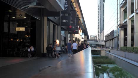 Canary-Wharf,-London,-United-Kingdom---August-2022---People-are-walking-on-the-walkway-next-to-a-small-canal-to-visit-shops-and-restaurant-in-the-corporate-district