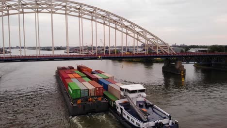 Drone-Shot-Of-Maas-Push-Tow-Barge-Carrying-Cargo-Containers-Along-River-Passing-Under-Bridge-Over-The-Noord