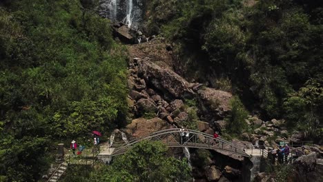 Una-Toma-Rápida-De-Un-Dron-Se-Invierte-Para-Revelar-Una-Transición-De-Una-Cascada-A-Turistas-Saludando-Desde-Un-Puente-Peatonal