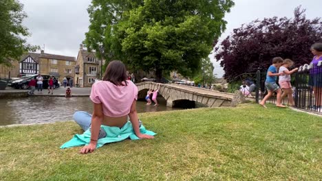 Chica-Caminando-Por-El-Camino-De-Cotswolds-Se-Relaja-En-La-Hierba-Junto-Al-Río-En-Bourton-On-The-Water,-Cotswolds---Inglaterra,-Reino-Unido