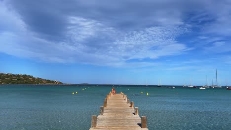 Tourists-on-wooden-pier-edge-of-Santa-Giulia-beach-on-Corsica-island-in-France-with-crystal-turquoise-water-of-Mediterranean-sea