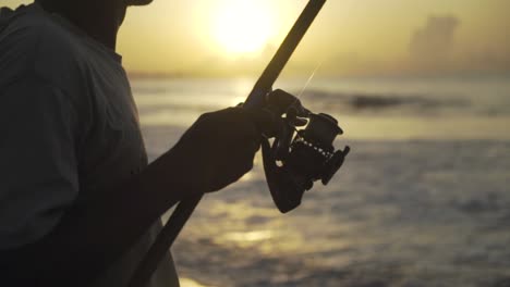 Close-up-on-the-fishing-rod-held-by-an-Indonesian-fisherman-fishing-on-a-coast-of-an-ocean-or-a-sea-at-Sunset-or-sunrise,-slow-motion
