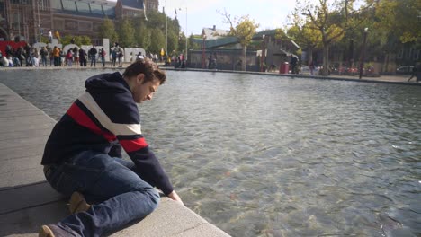 Apuesto-Joven-Sentado-En-La-Orilla-Del-Canal-Frente-Al-Rijksmuseum-Con-El-Cartel-I-Amsterdam-Poniendo-La-Mano-En-El-Agua