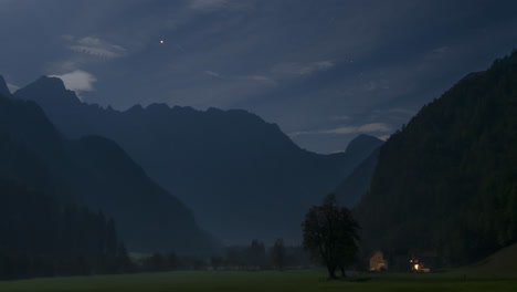 Timelapse-of-Alpine-valley-by-night-under-full-moon-light,-illuminated-farmhouse,-cottage,-full-moon,-clouds-and-stars-in-sky