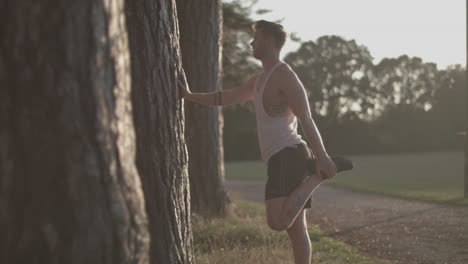 Man-In-Park-Stretching-Out-His-Legs-Before-He-Goes-For-a-Run-Whilst-Sun-Sets-Behind-Him-In-Slow-Motion---Ungraded