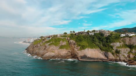 Full-panoramic-aerial-view-of-Joatinga-beach-in-Rio-de-Janeiro-with-its-cliff-rocks-at-high-tide,-a-small-beach,-the-Gavea-mountain-behind-it-and-in-the-distance-left-well-known-landmarks-of-the-city
