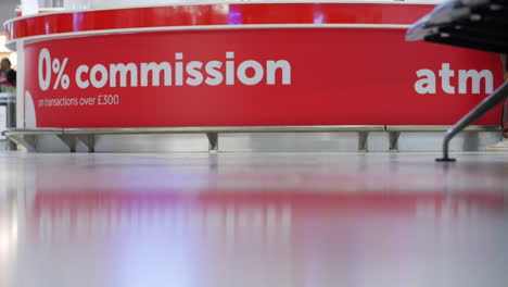 Currency-exchange-kiosk-with-passenger-legs-and-a-blue-rolling-suitcase